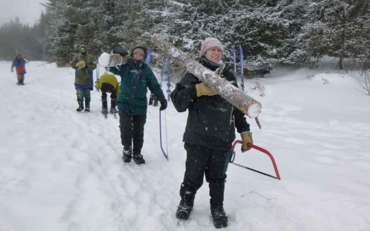 a group of gap year students carry a felled tree and saws across a snowy landscape